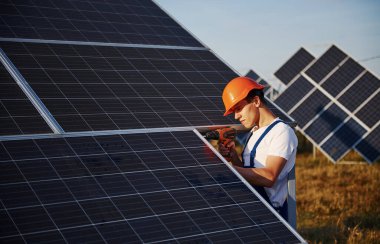 Kablosuz tornavida kullanıyor. Açık havada mavi üniformalı, güneşli bir günde güneş pilli bir erkek işçi..