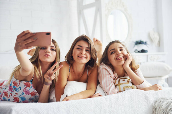 Making selfie by using pink phone. Happy female friends having good time at pajama party in the bedroom.