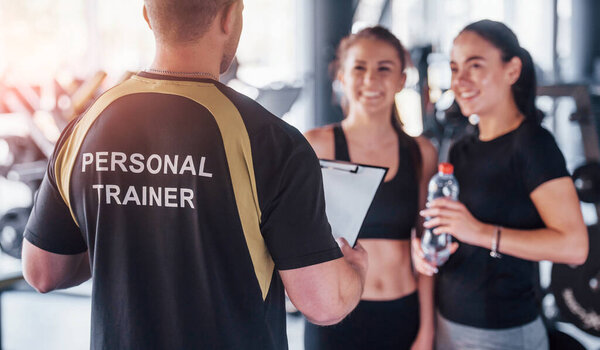 Two young girls in sportive clothes is in the gym together at daytime with their male personal trainer.