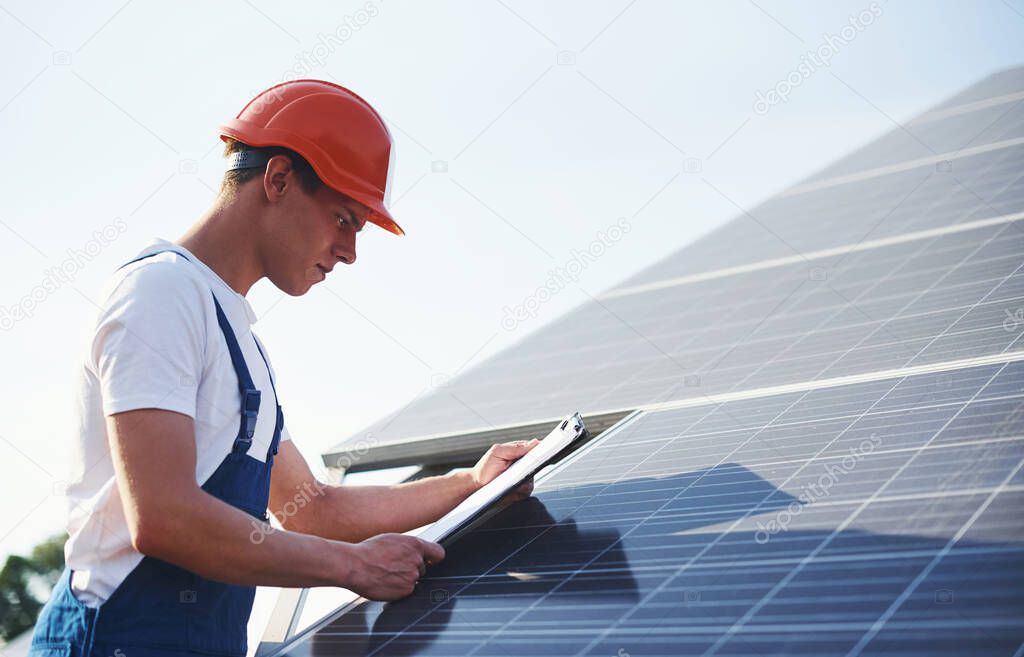 With documents. Male worker in blue uniform outdoors with solar batteries at sunny day.