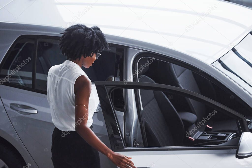 Young african american woman in glasses and with notepad stands in the car salon near vehicle.