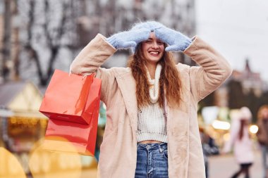 Elinde alışveriş poşetleri olan kalın elbiseli genç bir kız şehirde gezintiye çıkıyor..