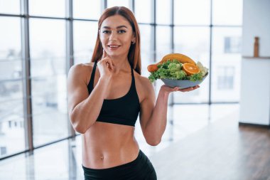 Neşeli sportif kadın elinde sağlıklı yiyeceklerle evde duruyor..