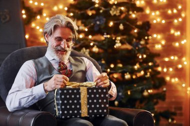 Gri saçlı ve sakallı son sınıf öğrencisinin portresi hediye kutusuyla süslenmiş Noel odasında..