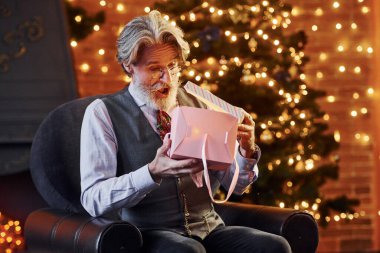 Gri saçlı ve sakallı son sınıf öğrencisinin portresi hediye kutusuyla süslenmiş Noel odasında..