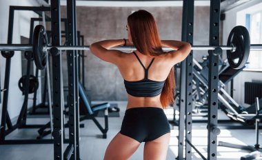 Halterin üzerine yaslan. Zayıf vücutlu ve siyah spor elbiseli genç fitness kadını spor salonunda..