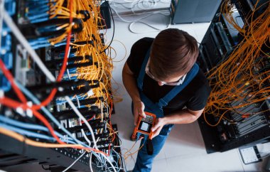 Sunucu odasında internet ekipmanı ve kablolarla çalışan ölçüm cihazıyla üniformalı genç adamın en iyi görüntüsü.