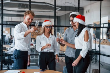 Resmi kıyafetli çalışanlar, şampanya bardaklı ve noel şapkalı işçiler ofiste yeni yılı kutluyorlar..