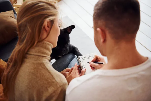 幸せな若い現代のカップルはソファに座ってメモ帳に願い事を書く彼らのかわいい犬と家で — ストック写真