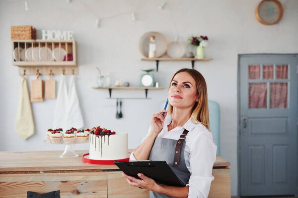 Woman with notepad in hands stands indoors at kitchen with cookies behind.