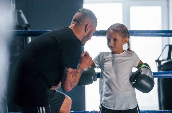 Standing on the boxing ring. Young tattooed boxing coach teaches cute little girl in the gym.