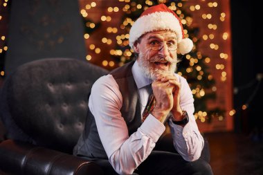 Süslü püslü, gri saçlı ve sakallı bir son sınıf öğrencisinin portresi süslü bir odada ve Noel şapkasıyla oturuyor..