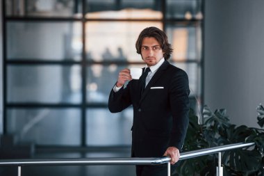 Siyah takım elbiseli ve kravatlı yakışıklı genç işadamının portresi bir bardak içkiyle içeride duruyor..