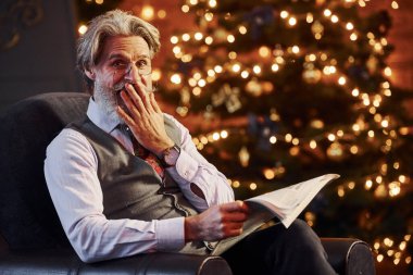 Gri saçlı ve sakallı son sınıf öğrencisinin portresi elinde kitapla süslenmiş Noel odasında..