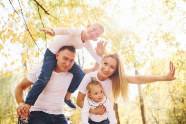 Anne ve baba çocukları omuzlarında ve ellerinde tutuyor. Neşeli genç aile birlikte bir sonbahar parkında yürüyüş yapıyor..