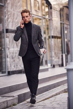 Elindeki not defteri ve içki bardağıyla şık giyinen adam dışarıda, modern binalara karşı. Telefonda konuşun..