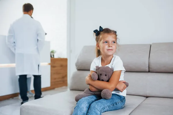 医生在后面可爱的小女孩手里拿着玩具熊坐在医院的候诊室里 — 图库照片