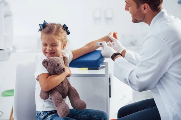 血液取样 带着玩具熊的小女孩和医生在诊所里 — 图库照片
