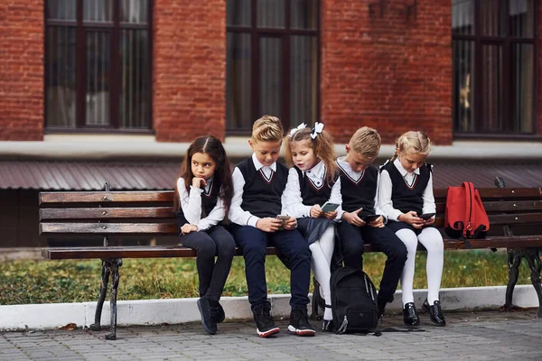 Grupa Dzieci Szkolnym Mundurku Siedzi Razem Ławce Pobliżu Budynku Edukacyjnego — Zdjęcie stockowe