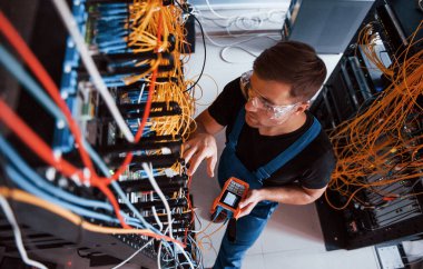 Sunucu odasında internet ekipmanı ve kablolarla çalışan ölçüm cihazıyla üniformalı genç adamın en iyi görüntüsü.