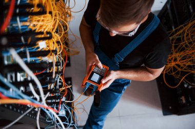 Sunucu odasında internet ekipmanı ve kablolarla çalışan ölçüm cihazıyla üniformalı genç adamın en iyi görüntüsü.