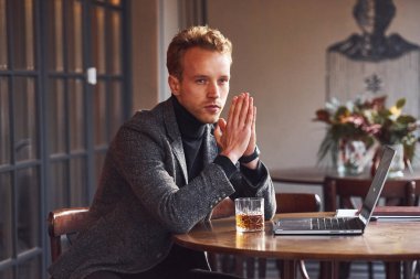 Resmi kıyafetli zarif bir genç, kafeteryada dizüstü bilgisayarı ve bir bardak alkolle oturuyor..