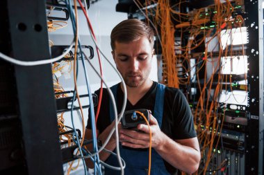 Ölçüm cihazlı üniformalı genç adam sunucu odasında internet ekipmanı ve kablolarla çalışıyor..
