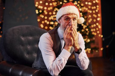 Süslü püslü, gri saçlı ve sakallı bir son sınıf öğrencisinin portresi süslü bir odada ve Noel şapkasıyla oturuyor..