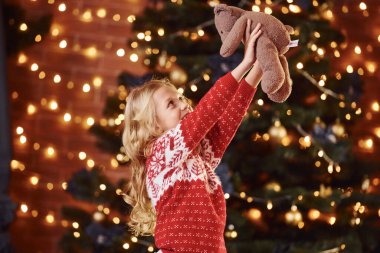 Kırmızı şenlikli kazak içinde oyuncak ayıyla yeni yılı ve Noel tatilini kutlayan tatlı küçük bir kız..