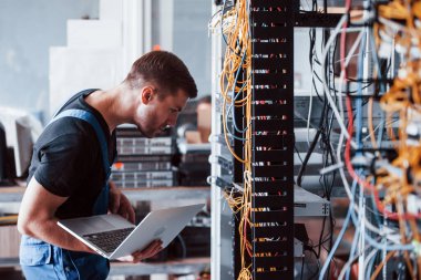 Üniformalı ve dizüstü bilgisayarlı genç adam sunucu odasında internet ekipmanı ve kablolarla çalışıyor..