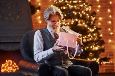 Gri saçlı ve sakallı son sınıf öğrencisinin portresi hediye kutusuyla süslenmiş Noel odasında..