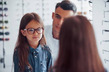 Beyaz önlüklü genç bir çocuk doktoru küçük kız için yeni gözlük almasına yardım ediyor..