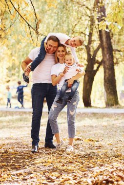 Anne ve baba çocukları omuzlarında ve ellerinde tutuyor. Neşeli genç aile birlikte bir sonbahar parkında yürüyüş yapıyor..