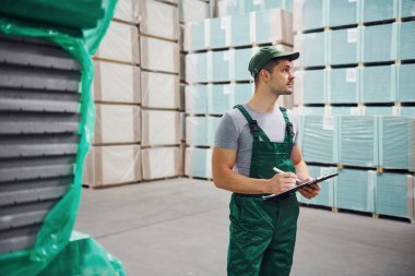 Yeşil üniformalı bir depo işçisi ve ellerindeki not defteri üretimi kontrol ediyor..