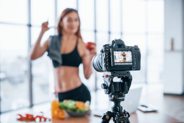 Spor vücutlu bir kadın vlogger 'ı. Sağlıklı yiyeceklerle birlikte evin içinde duruyor..