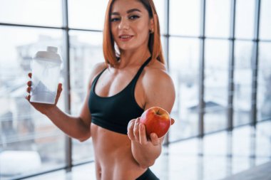 Güzel vücutlu sportif bir kadın elinde bir şişe su ve taze elmayla içeride duruyor..