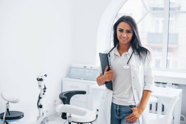 Elinde not defteriyle. Esmer kadın doktor modern ofiste duruyor ve kameraya bakıyor..