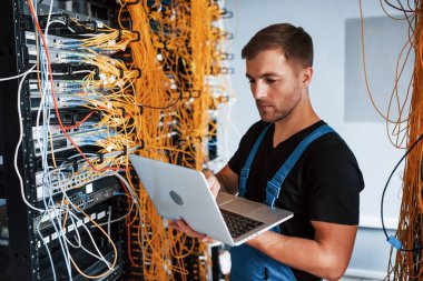Üniformalı ve dizüstü bilgisayarlı genç adam sunucu odasında internet ekipmanı ve kablolarla çalışıyor..