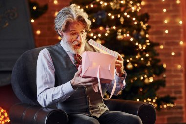 Gri saçlı ve sakallı son sınıf öğrencisinin portresi hediye kutusuyla süslenmiş Noel odasında..