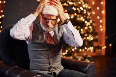 Süslü püslü, gri saçlı ve sakallı bir son sınıf öğrencisinin portresi süslü bir odada ve Noel şapkasıyla oturuyor..