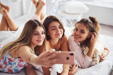 Pembe telefon kullanarak selfie çekerek. Mutlu bayan arkadaşlar yatak odasında pijama partisinde iyi vakit geçiriyorlar..