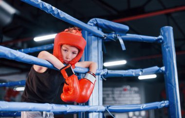 Boks ringinin düğümlerine dayanan koruyucu ekipmanlar içinde yorgun bir çocuk..