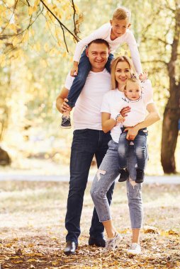 Anne ve baba çocukları omuzlarında ve ellerinde tutuyor. Neşeli genç aile birlikte bir sonbahar parkında yürüyüş yapıyor..