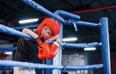 Boks ringinin düğümlerine dayanan koruyucu ekipmanlar içinde yorgun bir çocuk..