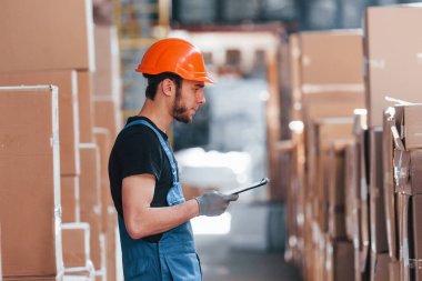 Üniformalı bir depo işçisi ve eldeki not defteri üretimi kontrol ediyor..