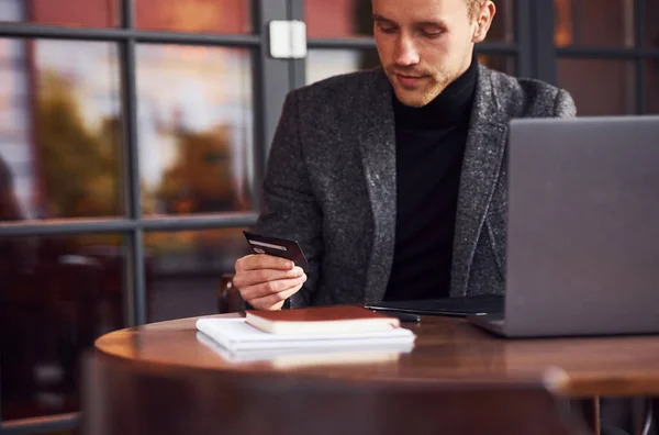 穿着正装的优雅年轻人手拿着笔记本电脑和信用卡坐在咖啡店里 — 图库照片