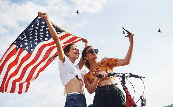 在多云的天空下 两个手拿着自行车和美国国旗的爱国快乐的女人自私自利 — 图库照片
