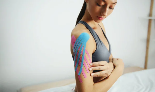 Sportive woman sits indoors with kinesio tape on her shoulder.