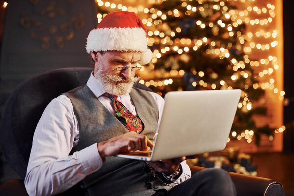 Portrait of stylish senior with grey hair and beard in decorated christmas room with laptop in hands.