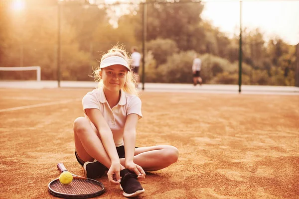 地面に座って 若い女性のテニス選手は スポーツウェアで屋外の裁判所にあります — ストック写真